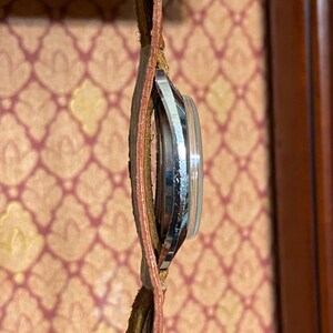 Może przedstawiać: Zegarek vintage z kopertą w kolorze srebrnym i brązowym sk&oacute;rzanym paskiem. Tarcza zegarka jest widoczna, a pasek wydaje się zużyty. Zegarek jest trzymany na tle wzorzystego tła.