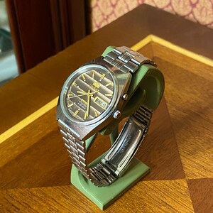 May include: A silver-toned Orient watch with a brown dial and a stainless steel band. The watch displays the day and date. The watch is displayed on a green stand, set against a wooden surface.