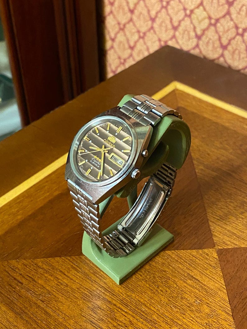 May include: A silver-toned Orient watch with a brown dial and a stainless steel band. The watch displays the day and date. The watch is displayed on a green stand, set against a wooden surface.