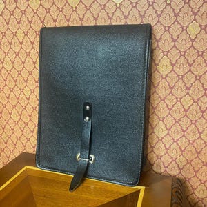Op de afbeelding: Een zwarte leren portfolio met een getextureerd oppervlak. Het heeft een riemsluiting met een metalen gesp en klinknagels. De achtergrond toont behang met patronen en een houten tafel.