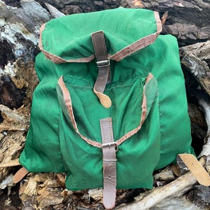 Op de afbeelding: Een groene rugzak met bruine leren riemen en accenten. De rugzak heeft twee voorzakken en een klepsluiting. De leren riemen hebben metalen gespen. De rugzak is gemaakt van een duurzame stof.