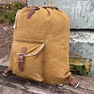 May include: A tan canvas backpack with a drawstring closure and a front pocket. The backpack features brown leather straps and silver buckles. The bag is sitting on a wooden surface.