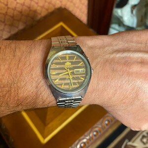 May include: A silver-toned Orient watch with a brown striped dial and a day-date display. The watch has a silver band and is worn on a wrist. The watch face has gold-colored accents and the word "Orient" is visible.