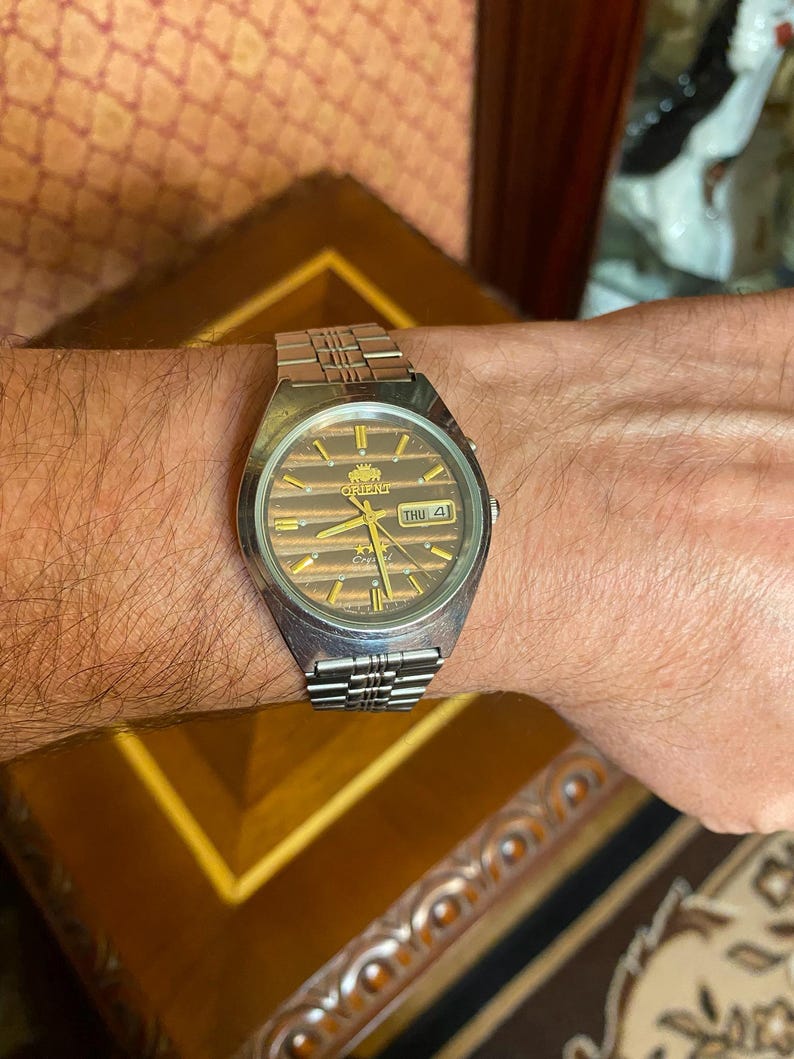 May include: A silver-toned Orient watch with a brown striped dial and a day-date display. The watch has a silver band and is worn on a wrist. The watch face has gold-colored accents and the word "Orient" is visible.