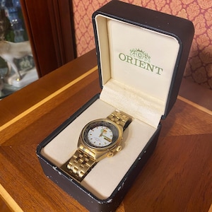 May include: A gold-toned Orient watch with a white face and gold band, displayed in a black box with the Orient logo. The watch features a detailed dial and is presented on a wooden surface.