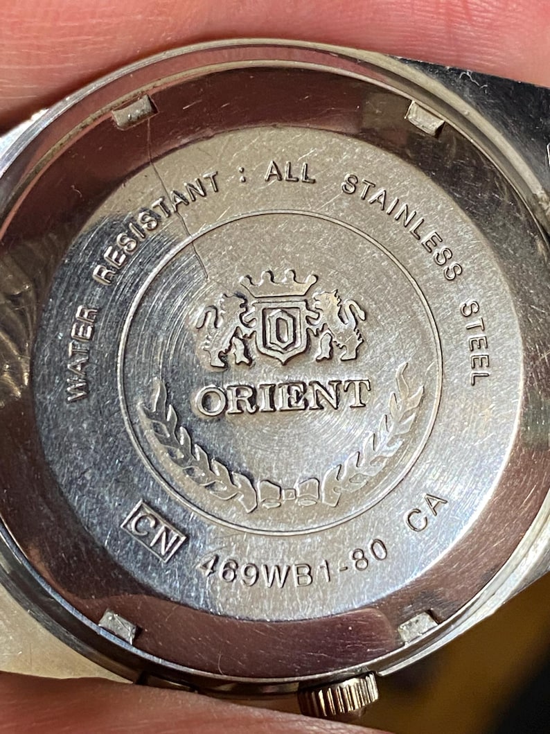 May include: Close-up of the back of a silver Orient watch. The watch back is engraved with the words "WATER RESISTANT", "ALL STAINLESS STEEL", and "ORIENT". The model number "469WB1-80 CA" is also visible.