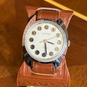 Może przedstawiać: Zegarek Vostok w stylu vintage ze srebrną kopertą i kremową tarczą. Zegarek posiada czarne cyfry i wskaz&oacute;wki oraz jest osadzony na brązowym sk&oacute;rzanym pasku. Na tarczy zegarka widnieje napis "Vostok".