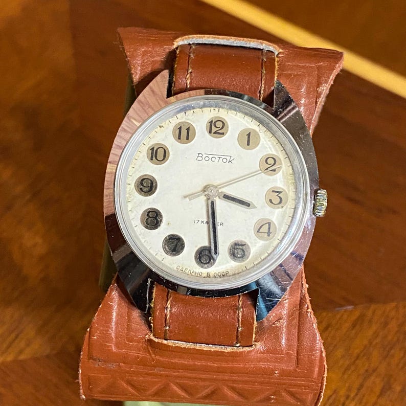 Może przedstawiać: Zegarek Vostok w stylu vintage ze srebrną kopertą i kremową tarczą. Zegarek posiada czarne cyfry i wskaz&oacute;wki oraz jest osadzony na brązowym sk&oacute;rzanym pasku. Na tarczy zegarka widnieje napis "Vostok".