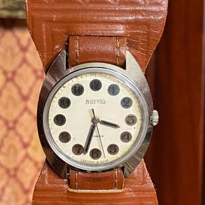 Może przedstawiać: Zegarek naręczny w stylu vintage ze srebrną kopertą i białą tarczą z czarnymi cyframi i wskaz&oacute;wkami. Zegarek ma brązowy sk&oacute;rzany pasek. Na tarczy zegarka widnieje napis "BOCTOK" i "CAEAAHO B CCCP".