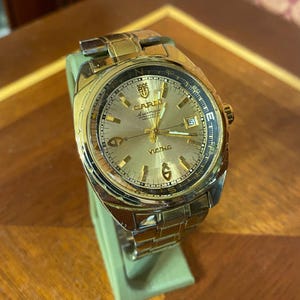 May include: A gold-tone automatic wristwatch with a metallic gold dial, featuring the text "CARNI" and "VICING" on the face. The watch includes a date window at the three o'clock position, gold hour markers, and a matching gold-tone metal link bracelet.
