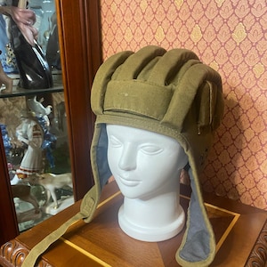 Puede incluir: Un casco de tanque militar verde oliva con un diseño acolchado. El casco tiene orejeras y una correa para la barbilla. Se exhibe sobre una cabeza de maniquí blanca contra un fondo estampado.