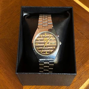 May include: A silver-toned Orient watch with a brown face and gold accents, displayed in a black box. The watch features a metal band and a day-date display. The watch face has the word "Orient" and "21 Jewels" printed on it.