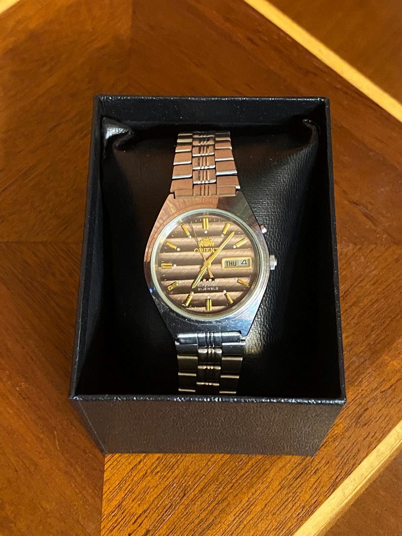 May include: A silver-toned Orient watch with a brown face and gold accents, displayed in a black box. The watch features a metal band and a day-date display. The watch face has the word "Orient" and "21 Jewels" printed on it.