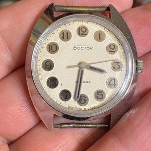 Może przedstawiać: Zegarek naręczny Vostok w stylu vintage ze srebrną kopertą i kremową tarczą. Zegarek posiada czarne cyfry, czarne wskaz&oacute;wki i napis "Vostok" na tarczy. Na tarczy znajduje się r&oacute;wnież napis "17 jewels" i "Made in USSR".