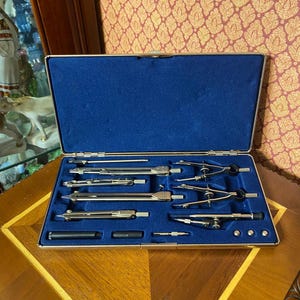 May include: A silver-toned metal drafting compass set housed in a rectangular case with a deep blue velvet interior. The set includes various precision drawing instruments, including dividers, compasses, and small metal attachments arranged in molded slots.
