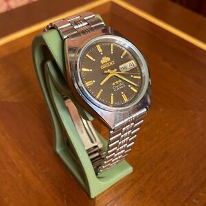 May include: A silver-toned Orient watch with a dark brown face and gold accents. The watch features a day and date display and is mounted on a green stand. The watch has a silver band.