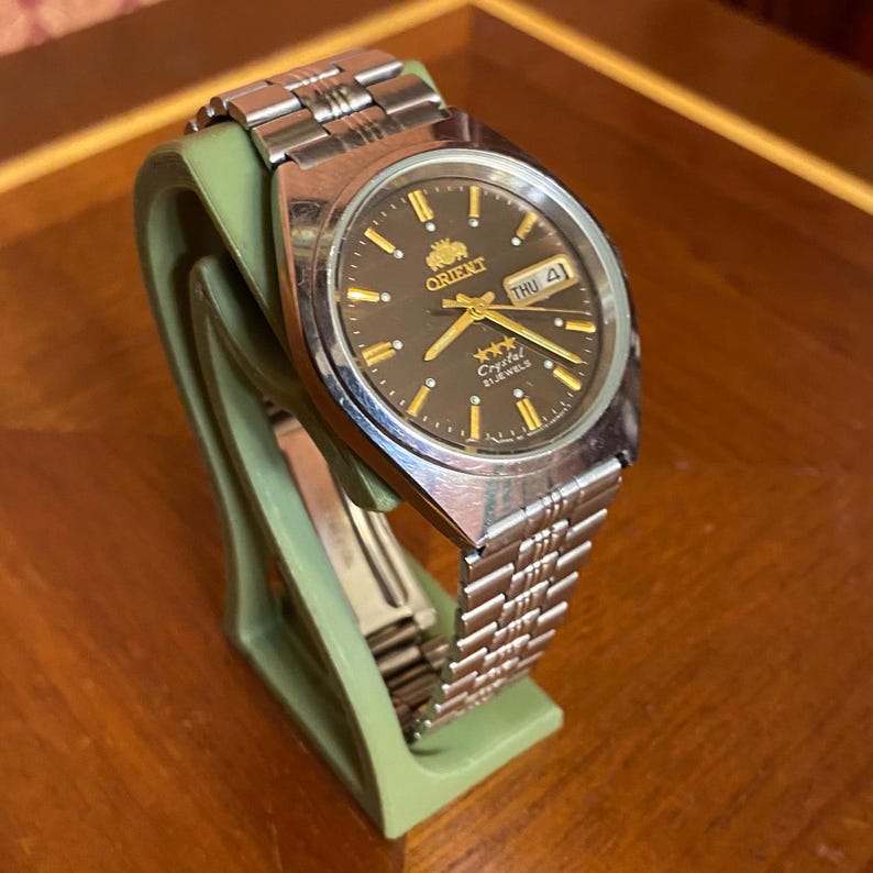 May include: A silver-toned Orient watch with a dark brown face and gold accents. The watch features a day and date display and is mounted on a green stand. The watch has a silver band.