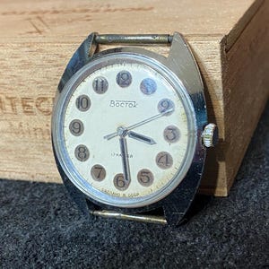 Może przedstawiać: Zegarek na rękę Vostok w stylu vintage ze srebrną kopertą i kremową tarczą. Zegarek posiada czarne cyfry, wskaz&oacute;wki i napis "Vostok" w cyrylicy. Zegarek leży na drewnianym pudełku i ciemnej tkaninie.
