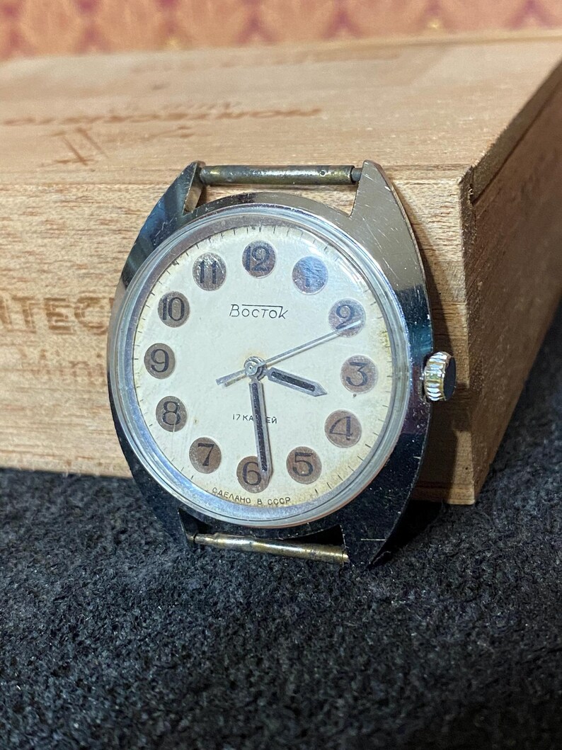 Może przedstawiać: Zegarek na rękę Vostok w stylu vintage ze srebrną kopertą i kremową tarczą. Zegarek posiada czarne cyfry, wskaz&oacute;wki i napis "Vostok" w cyrylicy. Zegarek leży na drewnianym pudełku i ciemnej tkaninie.
