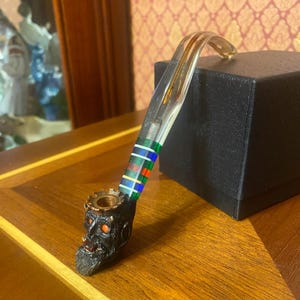May include: A decorative tobacco pipe featuring a clear, curved stem and a skull-shaped bowl. The stem is adorned with green, blue, and red stripes. The bowl is black with red eyes and a gold-coloured rim.