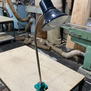 Op de afbeelding: Een zwarte metalen werklamp met een brede, conische kap en een witte binnenkant. De lamp heeft een flexibele, zilverkleurige zwanenhalsarm bevestigd aan een vierkante, groene voet met een kleine tuimelschakelaar en een wit netsnoer.