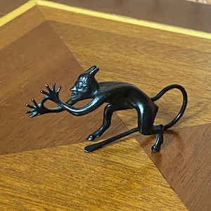 Op de afbeelding: Een donker, gestileerd beeldje van een duivelachtig wezen met uitgestrekte armen en een lange staart. De sculptuur heeft een gedetailleerd gezicht en staat in een dynamische, rennende positie. Het beeldje is gemaakt van donker metaal en staat op een houten oppervlak.