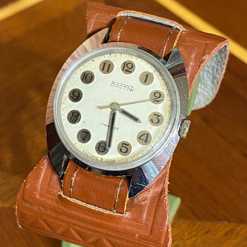 Może przedstawiać: Zegarek naręczny w stylu vintage z białą tarczą, czarnymi cyframi i srebrnymi wskaz&oacute;wkami. Na tarczy widnieje napis "Восток" i "17 камней". Osadzony w srebrnej kopercie i przymocowany do szerokiego, brązowego sk&oacute;rzanego paska.
