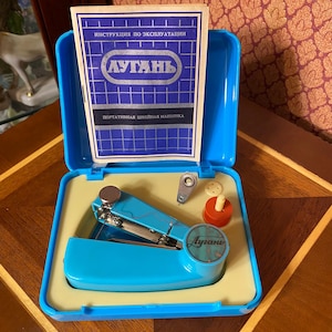May include: A vintage blue portable sewing machine kit. The kit includes a small sewing machine, a needle threader, a spool of thread, and a booklet. The sewing machine is in a blue plastic case with a yellow interior.