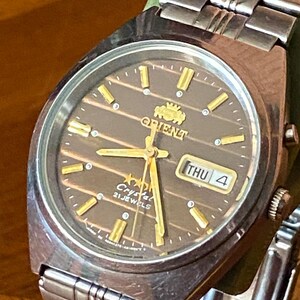 May include: A vintage Orient Crystal 21 Jewels wristwatch. The watch features a brown dial with gold-colored hands and markers, and a day-date display. The watch has a stainless steel band and a green strap. The watch face reads "Orient Crystal 21 Jewels".