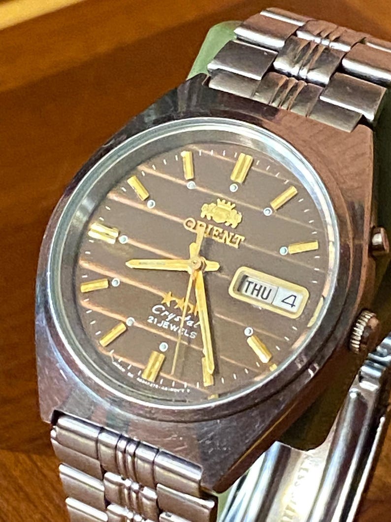 May include: A vintage Orient Crystal 21 Jewels wristwatch. The watch features a brown dial with gold-colored hands and markers, and a day-date display. The watch has a stainless steel band and a green strap. The watch face reads "Orient Crystal 21 Jewels".
