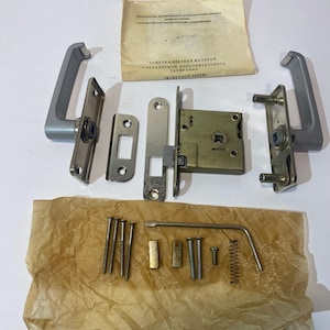 May include: A set of vintage door hardware, including two silver handles, a latch mechanism, a strike plate, and various screws and components. A paper with text is also included. The hardware is arranged on a brown paper.
