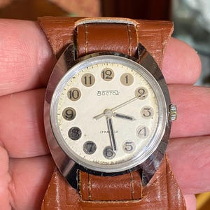 Może przedstawiać: Zegarek na rękę Vostok w stylu vintage ze srebrną kopertą i kremową tarczą. Zegarek posiada czarne cyfry, czarne wskaz&oacute;wki i napis "Vostok" na tarczy. Jest przymocowany do brązowego sk&oacute;rzanego paska.
