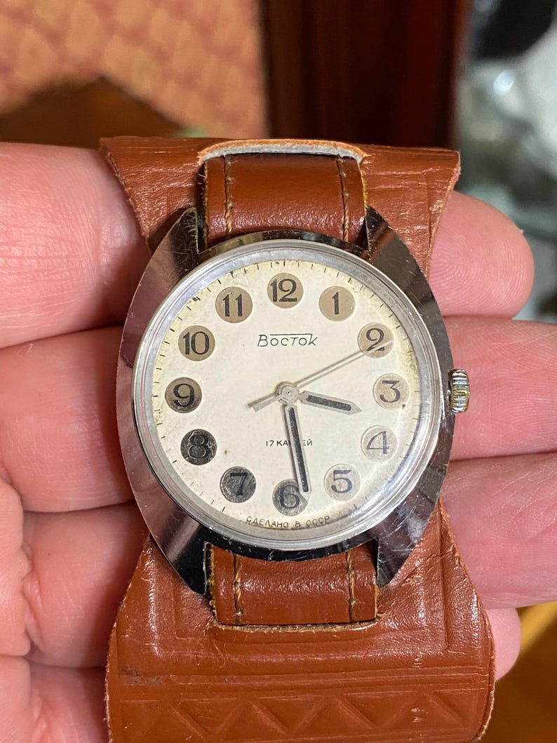 Może przedstawiać: Zegarek na rękę Vostok w stylu vintage ze srebrną kopertą i kremową tarczą. Zegarek posiada czarne cyfry, czarne wskaz&oacute;wki i napis "Vostok" na tarczy. Jest przymocowany do brązowego sk&oacute;rzanego paska.