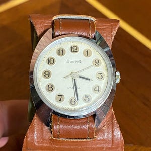 Może przedstawiać: Zegarek na rękę Vostok w stylu vintage z kremową tarczą, srebrną ramką i brązowym sk&oacute;rzanym paskiem. Tarcza zegarka wyświetla czarne cyfry i słowo "Восток". Zegarek jest umieszczony w brązowym sk&oacute;rzanym etui.