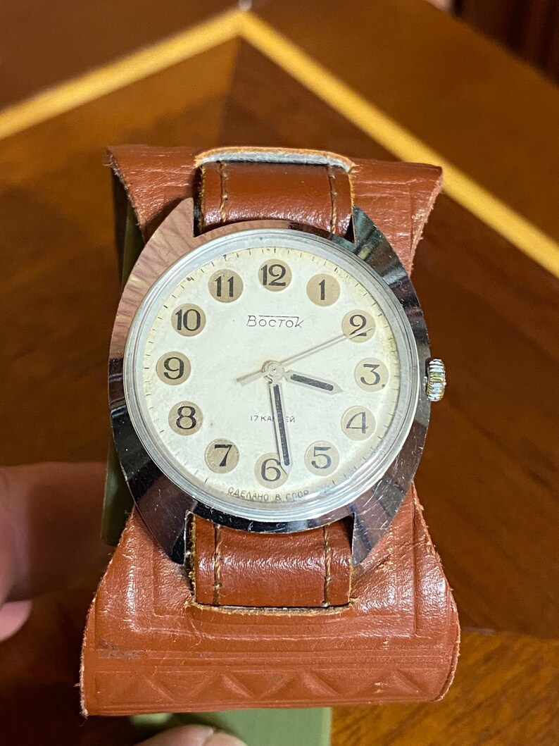 Może przedstawiać: Zegarek na rękę Vostok w stylu vintage z kremową tarczą, srebrną ramką i brązowym sk&oacute;rzanym paskiem. Tarcza zegarka wyświetla czarne cyfry i słowo "Восток". Zegarek jest umieszczony w brązowym sk&oacute;rzanym etui.