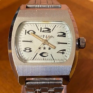 May include: A silver-toned wristwatch with a rectangular case and a textured metal link bracelet. The off-white dial features black Arabic numerals at 12, 10, 8, 6, 4, and 2, with three small stars below the center and the text ORNAVIN.