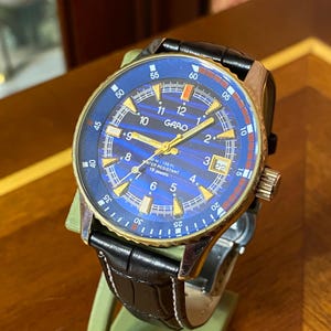 May include: A gold-toned watch with a blue face and black leather strap. The watch face displays gold numerals and hands, with the brand name "GARO". The watch is water resistant and has 19 jewels.