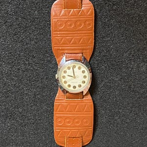 Może przedstawiać: Zegarek vintage z brązowym sk&oacute;rzanym paskiem w stylu mankietu. Tarcza zegarka jest srebrna z czarnymi cyframi i wskaz&oacute;wkami. Pasek posiada geometryczne wzory i zapięcie na sprzączkę. Klasyczny dodatek.