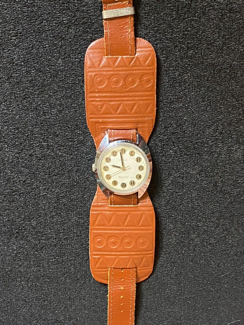 Może przedstawiać: Zegarek vintage z brązowym sk&oacute;rzanym paskiem w stylu mankietu. Tarcza zegarka jest srebrna z czarnymi cyframi i wskaz&oacute;wkami. Pasek posiada geometryczne wzory i zapięcie na sprzączkę. Klasyczny dodatek.