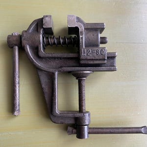 May include: A vintage, dark gray metal vise with a screw mechanism and a handle. The vise has a spring and the number "42-80" is stamped on the side. It is designed to clamp and hold objects securely.