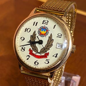 Peut inclure: Une montre-bracelet dorée avec un cadran crème, des chiffres arabes noirs et un bracelet en maille métallique. Le cadran présente un emblème central avec une étoile rouge, une couronne de laurier et une bannière rouge affichant 1945 et 1995. Un guichet de date est à trois heures, et le mot РОССИЯ est inscrit en bas.