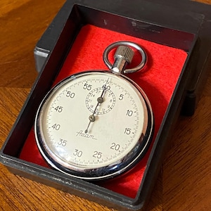 May include: A vintage stopwatch with a silver-colored case and a white face, displayed in a black box with a red velvet lining. The stopwatch has a small dial in the center and the brand name "Azam" is printed on the face.
