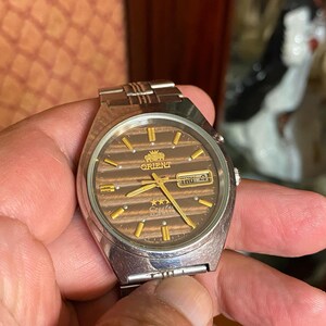 May include: A vintage Orient watch with a brown dial and gold accents. The watch face displays the day and date. The watch has a silver-tone metal band and case. The watch is being held by a person.