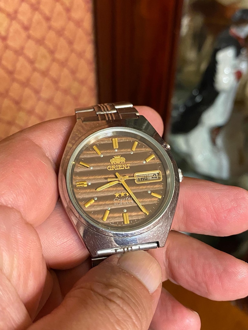 May include: A vintage Orient watch with a brown dial and gold accents. The watch face displays the day and date. The watch has a silver-tone metal band and case. The watch is being held by a person.
