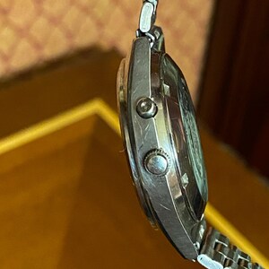 May include: A silver-toned wristwatch with a metal band. The watch face is visible, and the side profile shows two buttons. The watch is attached to a silver metal band.