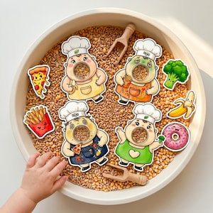 May include: A sensory bin with wheat kernels, featuring chef-themed character cutouts with food items like pizza, fries, and a banana. Includes a wooden scoop and a child's hand reaching in.