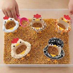 Puede incluir: Un recipiente de plástico transparente lleno de granos, con cinco recipientes decorativos con forma de pollo. Cada pollo tiene una cresta roja, un pico amarillo y patas naranjas. Una cuchara rosa vierte granos en uno de los recipientes.