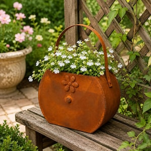 May include: A rust-coloured metal planter shaped like a handbag with a curved handle and a small flower-shaped relief on the front. The planter is filled with small white flowers and sits on a wooden surface, showcasing a weathered finish.
