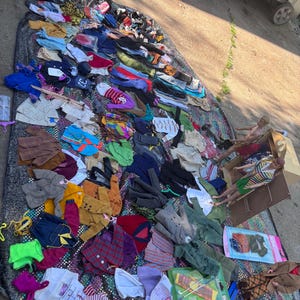 May include: A variety of children's clothing items are displayed on a patterned blanket. The assortment includes shirts, pants, and jackets in various colors and patterns. Some items are neatly folded, while others are scattered, suggesting a sale or display.