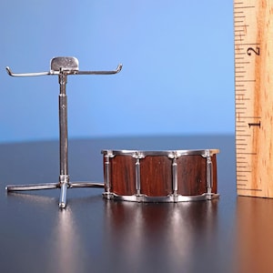 May include: A silver drum stand with a wooden drum on a black surface. The drum is about 5 inches in diameter and has a brown wood finish.